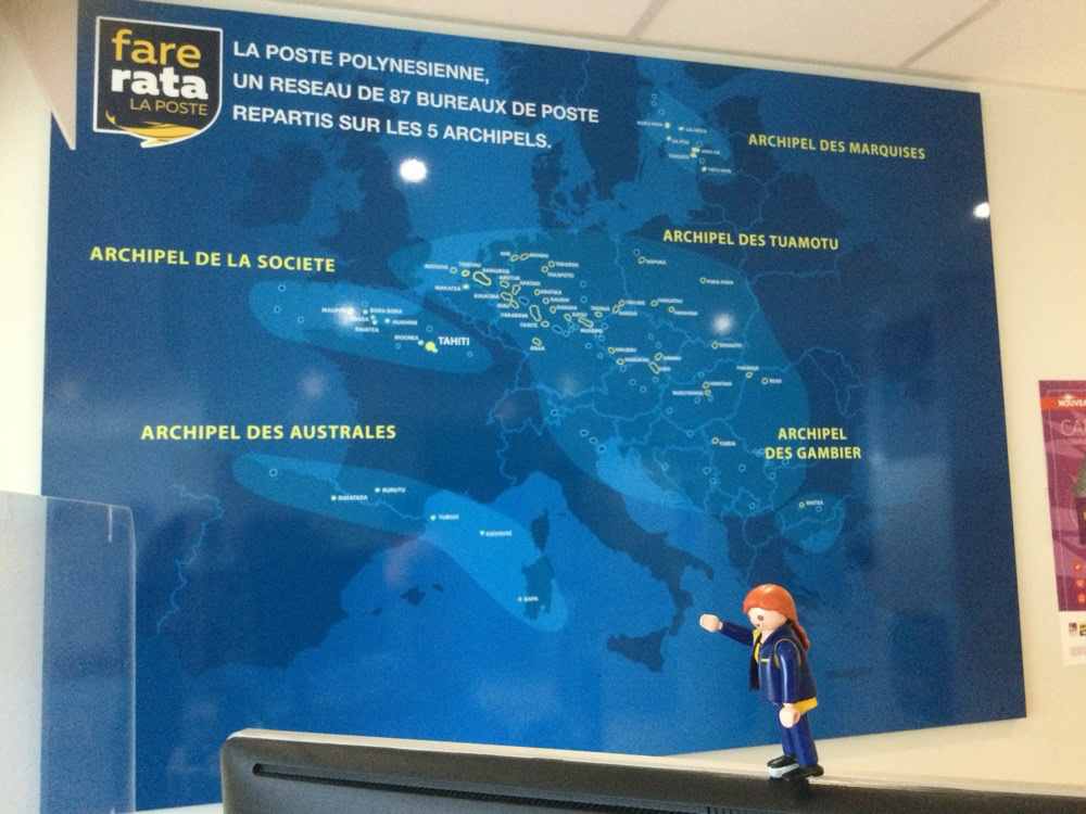 The Little Mail Carrier figure standing in front of a large blue map of French Polynesia’s postal network, showing the Society, Tuamotu, Marquesas, Austral and Gambier archipelagos.
