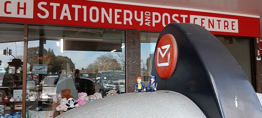 Two photos: staff hand-cancels postcards at a NZ post shop; a Gandalf statue stands beside a barrel-style mailbox indoors.
