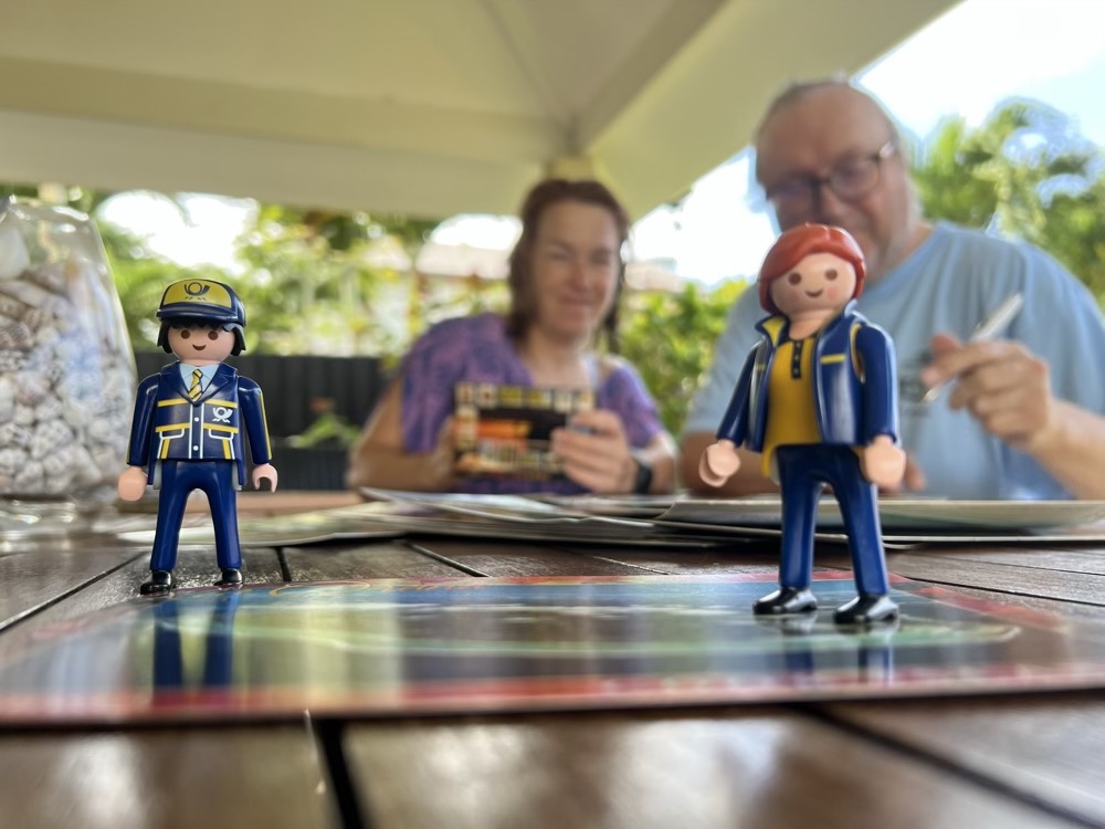 The Little Mail Carrier figures standing on a table with postcards, with their hosts Maret and Andry writing cards in the background.