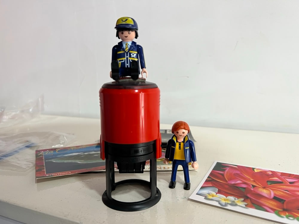 The Little Mail Carrier figures beside a large red postmark stamp and postcards showing Cook Islands flowers.
