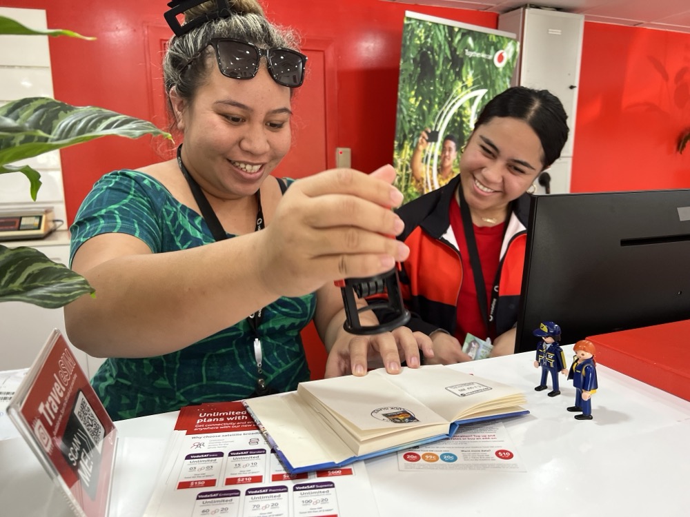 Two smiling post office staff members hand-cancelling postcards at the counter, with the Little Mail Carrier figures standing nearby.