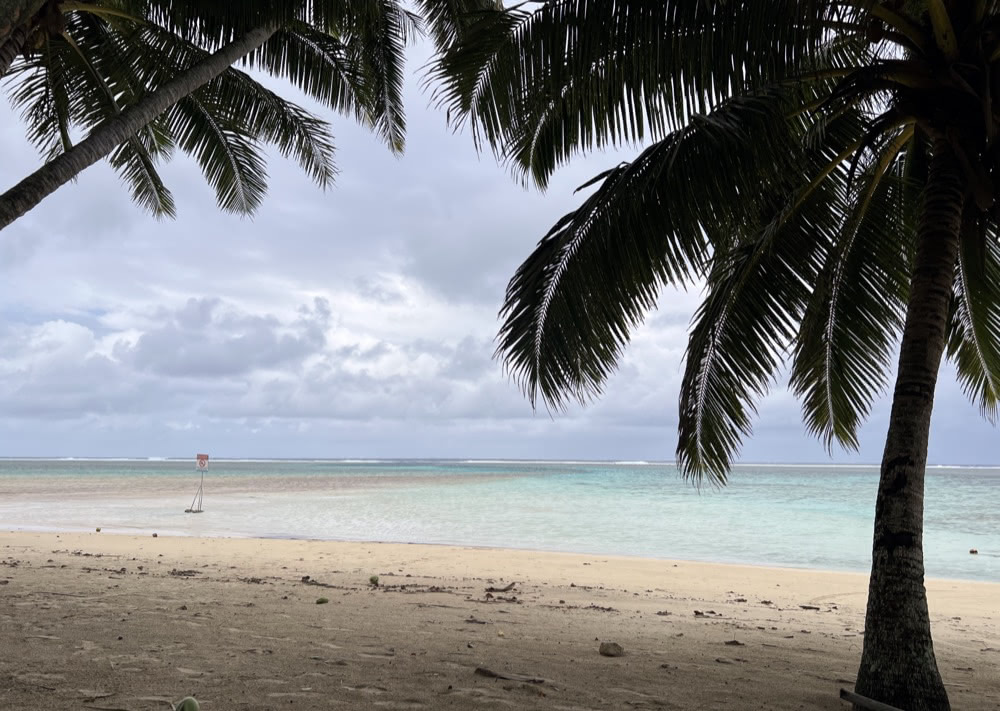 Rarotonga beach framed by palm trees, with pale sand, turquoise lagoon water and cloudy sky in the distance.