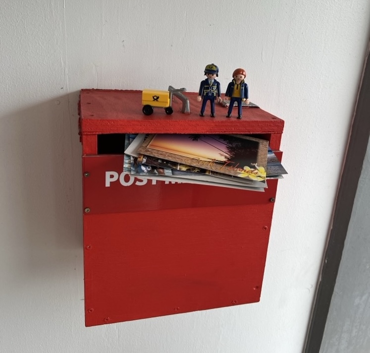 The Little Mail Carrier figures and their yellow mail cart standing on top of a red wall-mounted post box full of postcards.