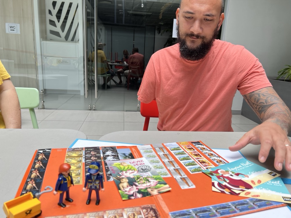 A man sitting at a table with the Little Mail Carrier figures, postcards, colourful French Polynesia stamps and a local Father Christmas postcard.