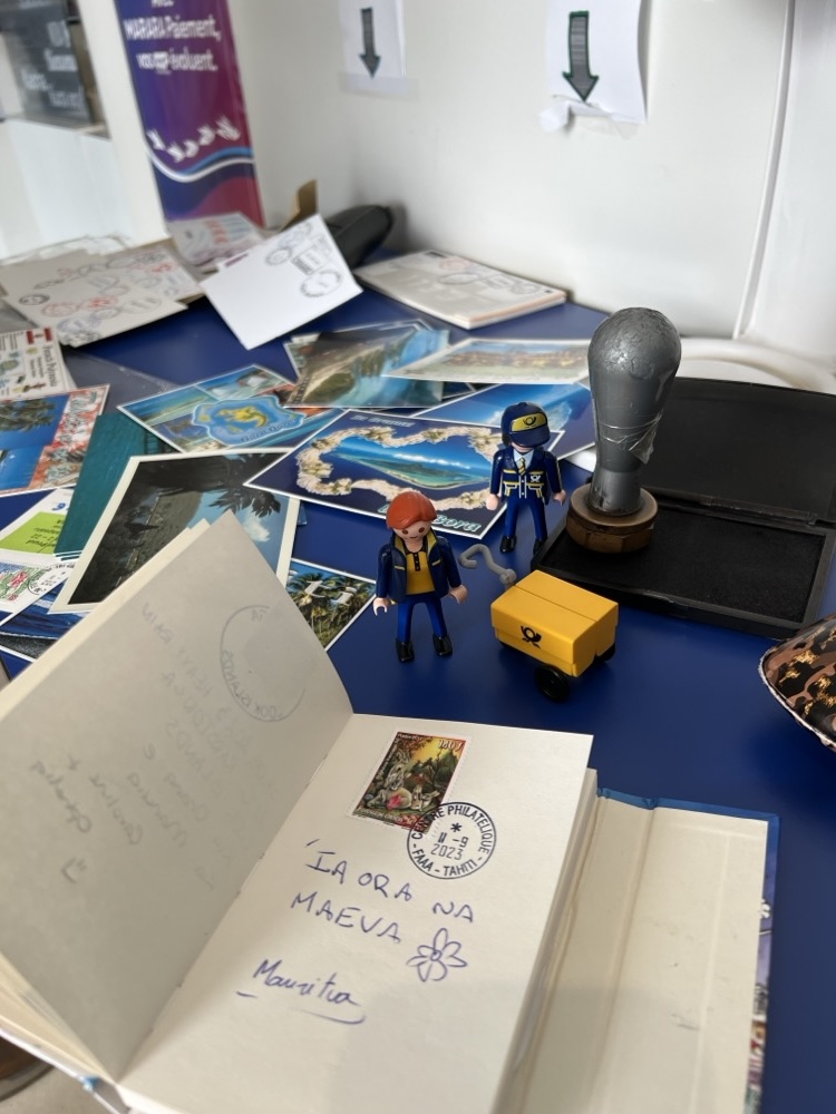 The Little Mail Carrier figures and their yellow mail cart beside a large postmark stamp, with postcards and a stamped notebook page on the table.