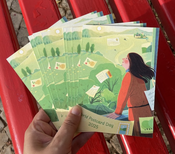 A hand holding several copies of the official World Postcard Day 2025 postcard in front of a bright red postbox. The postcards feature an illustration of a person standing in a green landscape, watching postcards float through the sky.