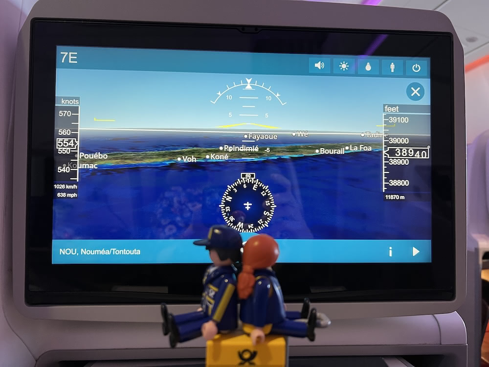 The toy postal carriers sit on top of a yellow mail trolley in front of an in-flight screen showing the plane’s location near Nouméa.