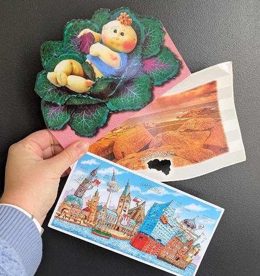 Rebecca holds up her three postcards: at the top the Cabbage Patch kids card, then a view card from Bretagne with a leaf-shaped cutout in it, and one from Germany with art of buildings that have faces and funny expressions