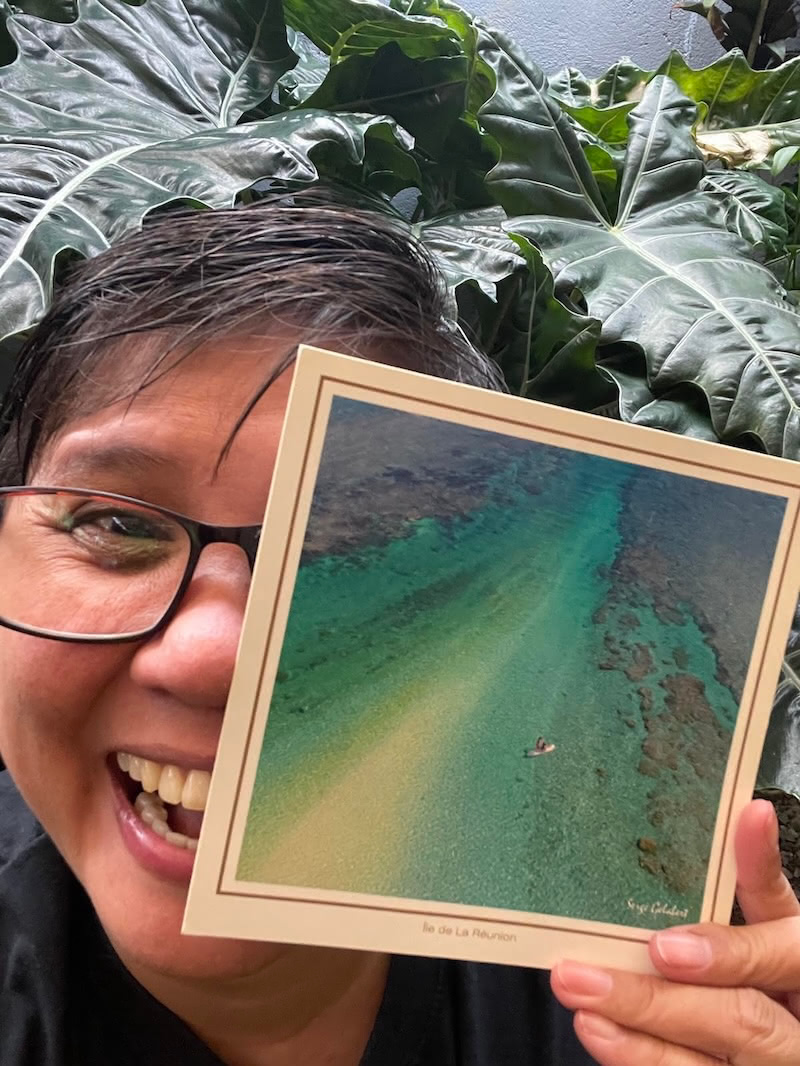 A smiling LeilaKewl holds a postcard from Réunion, showing a coral reef