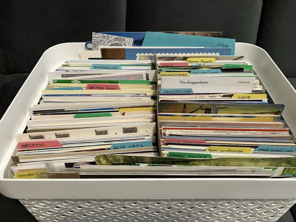 A white basket full of postcards sorted into labeled categories such as Postcrossing, architecture, cute, Harry Potter and British Post.