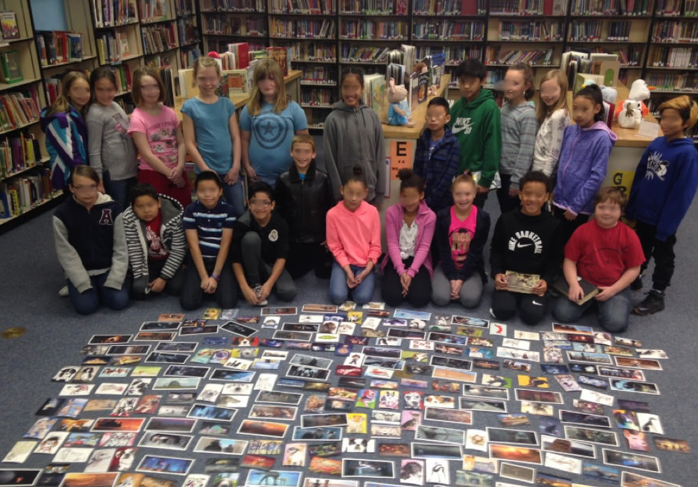 A group of schoolchildren in a library posing behind many postcards spread out on the floor.