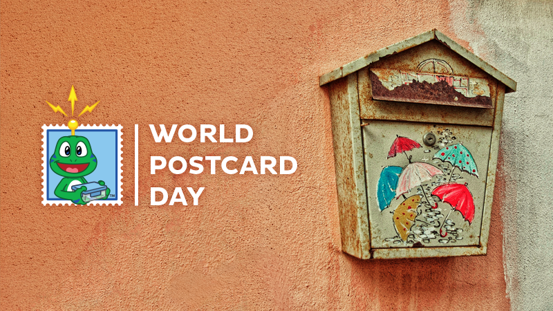 World Postcard Day graphic with the Geocaching frog logo beside the words “World Postcard Day”, next to a weathered mailbox painted with colorful umbrellas on an orange wall.