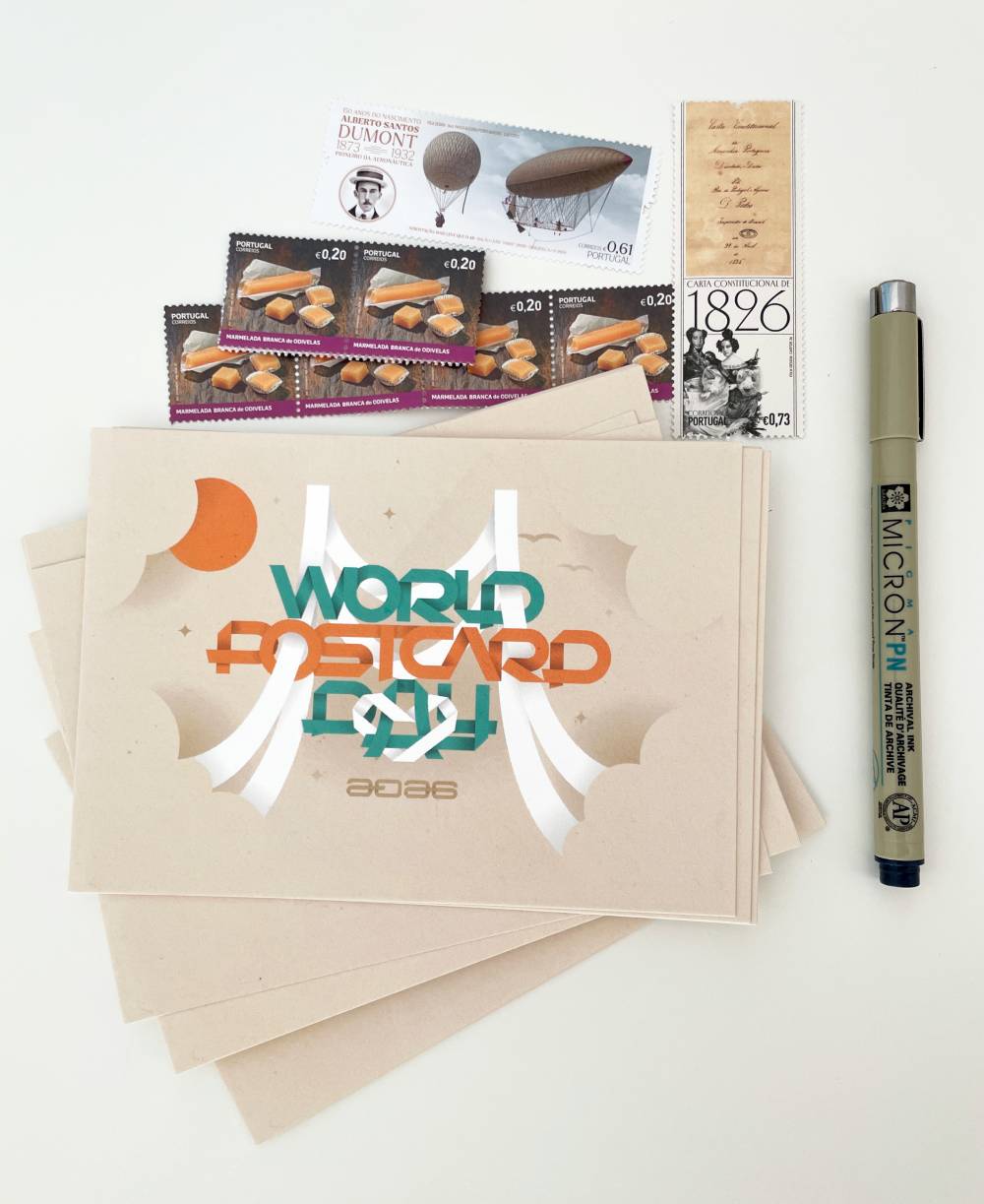 A bunch of World Postcard Day cards lie on a desk, surrounded by a pen and a few postage stamps 