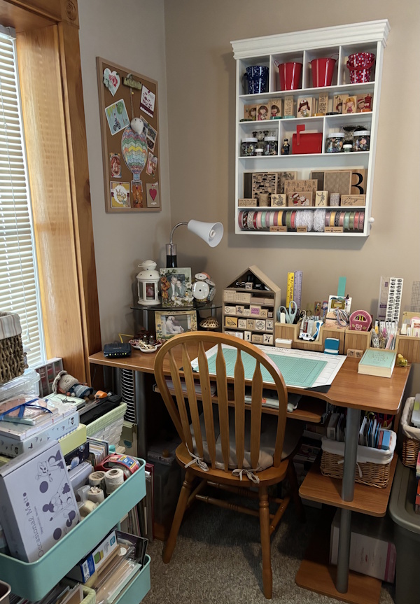 A wooden desk near a window, with a wall cabinet above full of various crafting supplies, a trolley with trays just behind, and other organisers holding other supplies on the desk and shelves