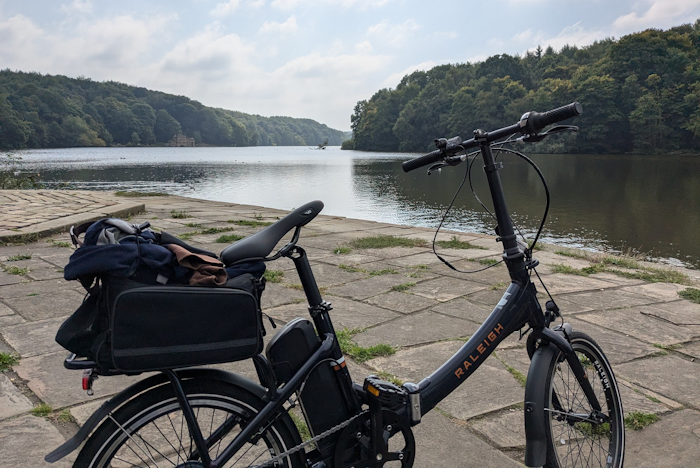 A photo of a bike parked on its stand in front of a lake, with a blue sky above and trees all around the lake