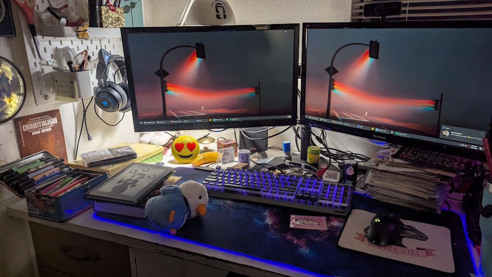 A desk with two PC monitors set up, but also plenty of space to write postcards, and various supplies like sharpies and postcards to hand