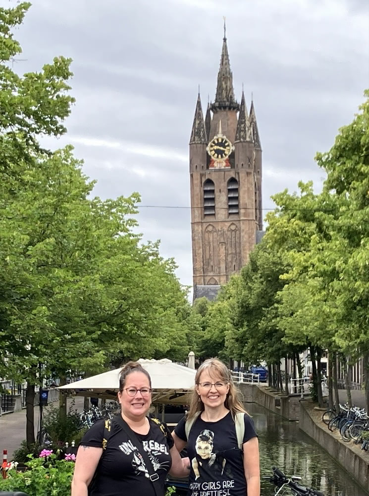 Two postcrossers smiling together by a canal in Delft, with a tall church tower in the background.