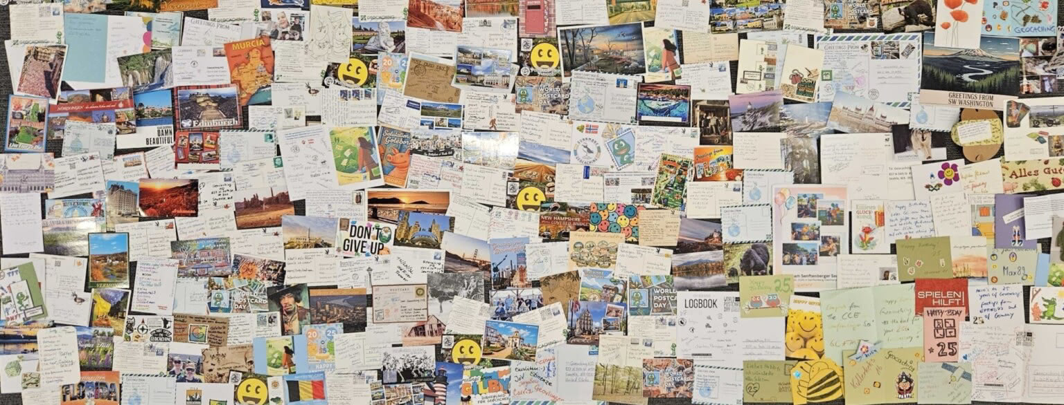 Large collage of postcards and handwritten cards covering a wall at Geocaching HQ, including World Postcard Day designs, travel postcards and Geocaching-themed messages.