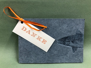 One of Lubach's print cards, which is grey and shows a fish outlined on it with some detail. A ribbon attaches a little card saying danke