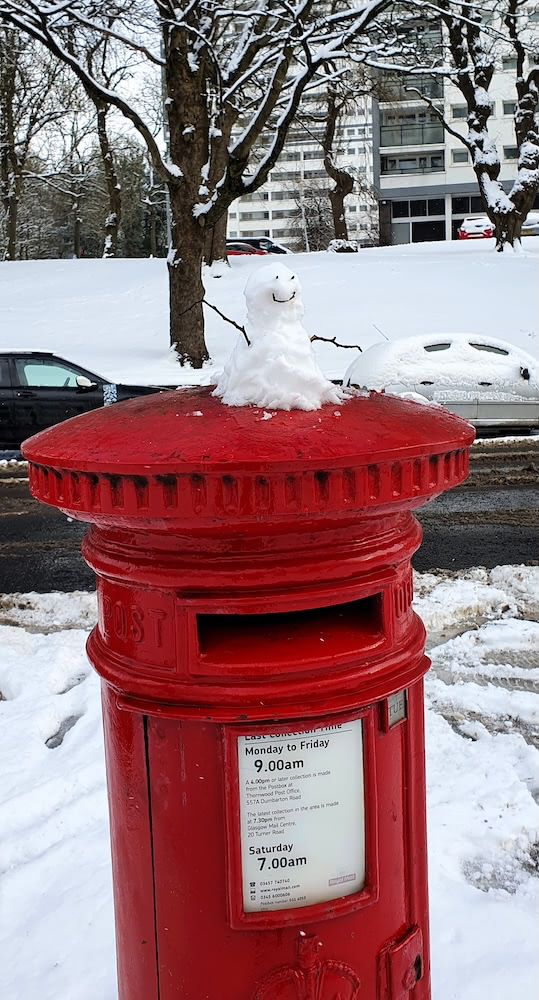 A classic red British postbox with a tiny snowman sitting on top, surrounded by snowy trees.
