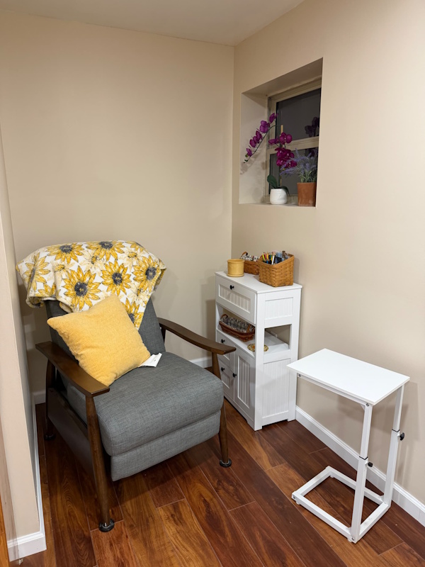 A comfy armchair with a sunflower blanket and yellow cushion, next to a window with flowers on the sill and a white cabinet with writing supplies, and a small table which can be pulled over for writing