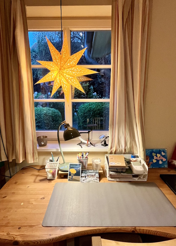 A wooden desk in front of a window, looking bright and cosy with some supplies neatly organised in front of a desk mat, and a glowing yellow star hung in the window
