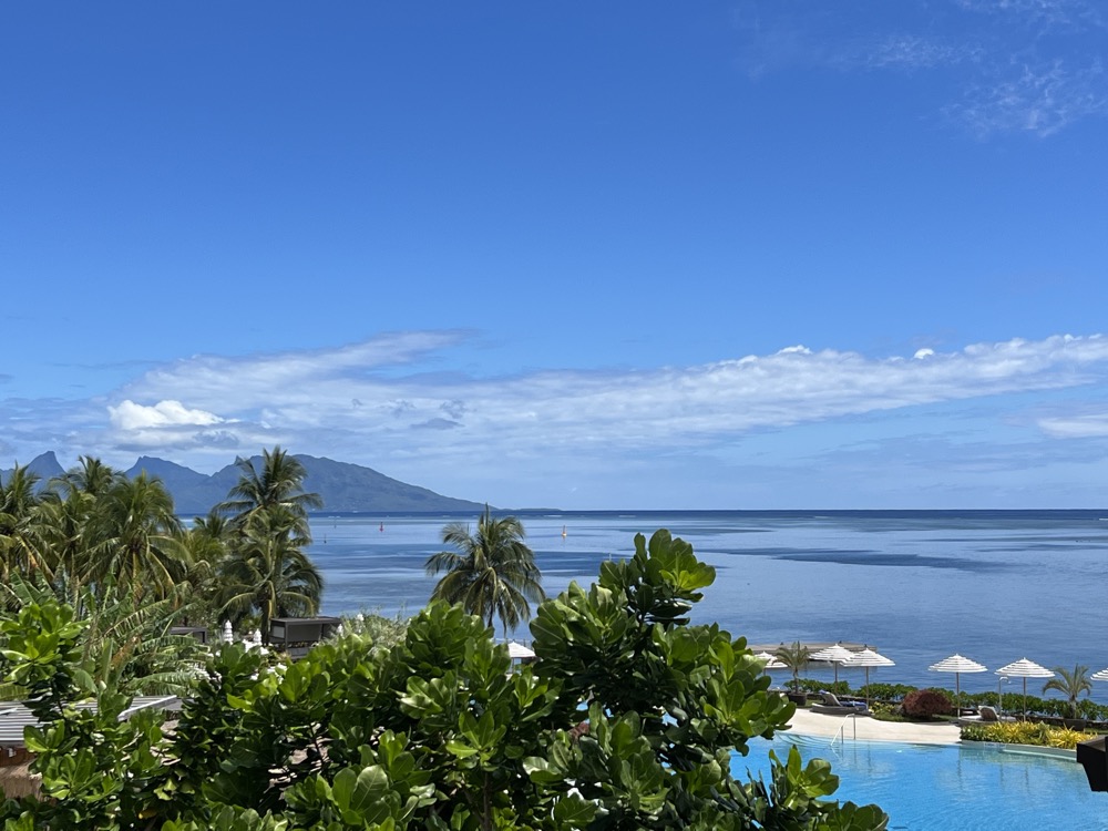 View over a blue lagoon in Tahiti, with palm trees, mountains and a swimming pool in the foreground.