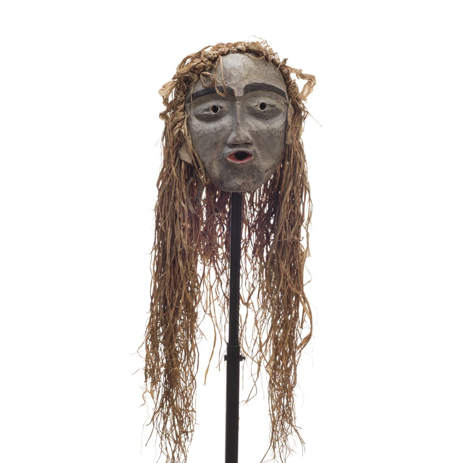 A small forest spirit mask, mostly white face, pursed red lips, prominent black eyebrows, long cedar trim draping from top and back