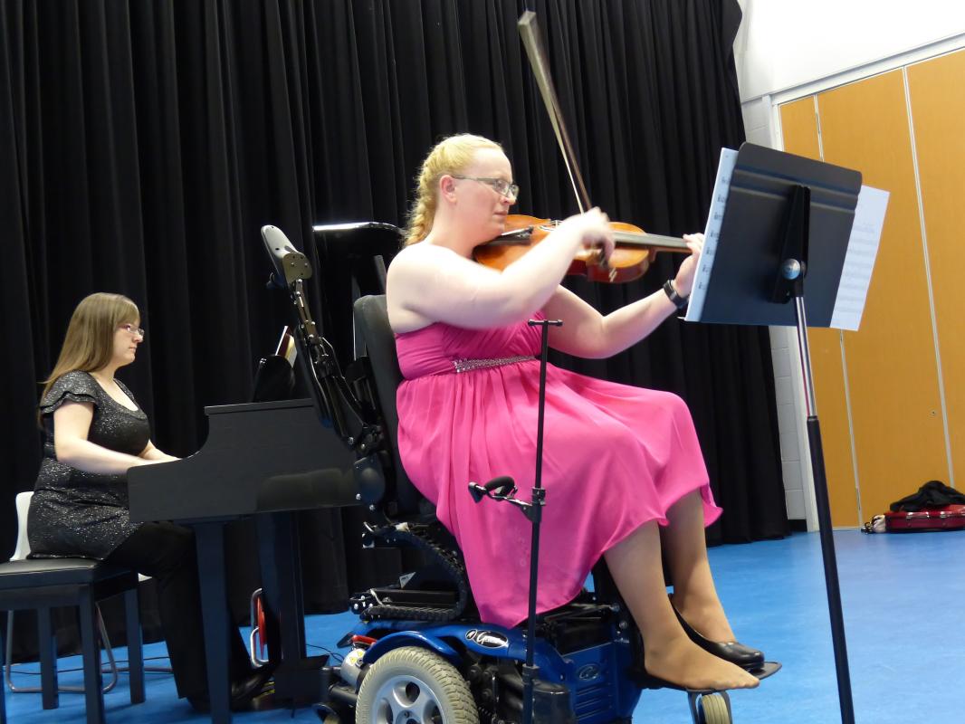 A blonde lady in a powered wheelchair plays a viola with a pianist