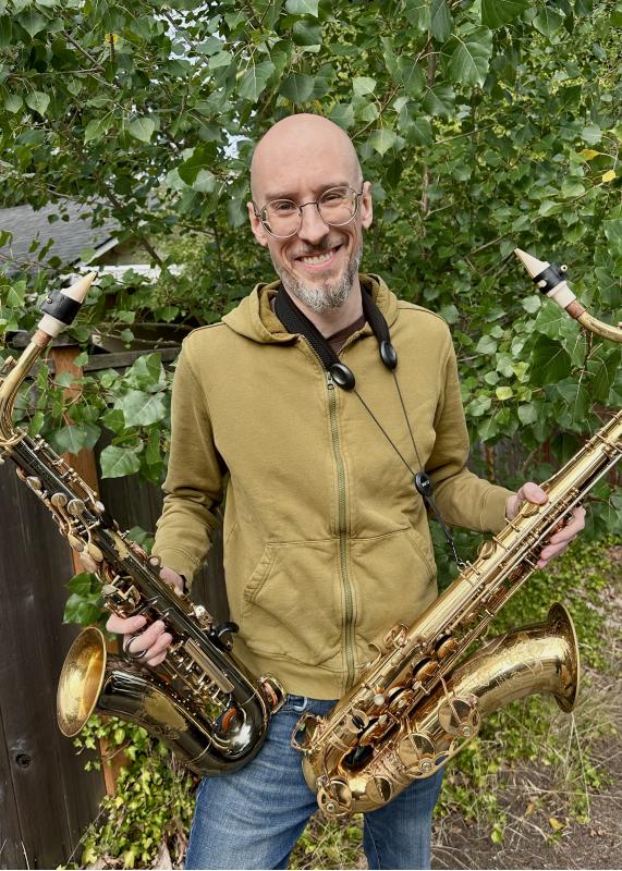 Middle aged white man, bald with glasses and a short beard, smiling and standing in front of a poplar tree. He is holding two saxophones, tenor in right hand and alto in the left.