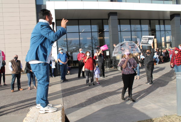 A man stands on a stone bench holding a microphone, and in front of protestors at Lubbock's Citizens Tower building.