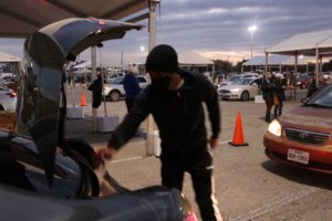 a person wearing a face mask loading food bank boxes into a car trunk in a busy parking lot