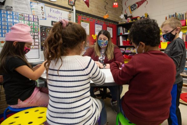 first grad studnets sit around a table with their teacher. One is a girl wearing a pinkt winter hat, anothe girl wears a white sweater with blue stripes and a boy wears a red sweatshirt