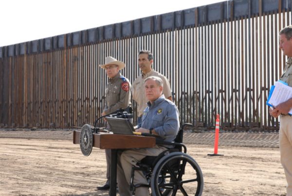 a man in a blue shirt and sitting in a wheelchair at a lectern in front of a tall metal fence with police and other officials around him