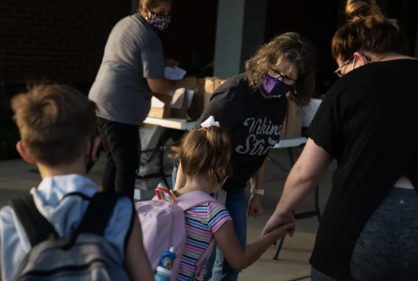 Two adults hold the hand of a young girl wearing a backpack to welcome her on the first day of school