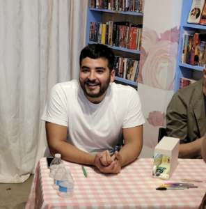 A photo of the author at a book signing event. He's wearing a white t-shirt and smiling while sitting at a table covered in a pink and white checked table cloth.