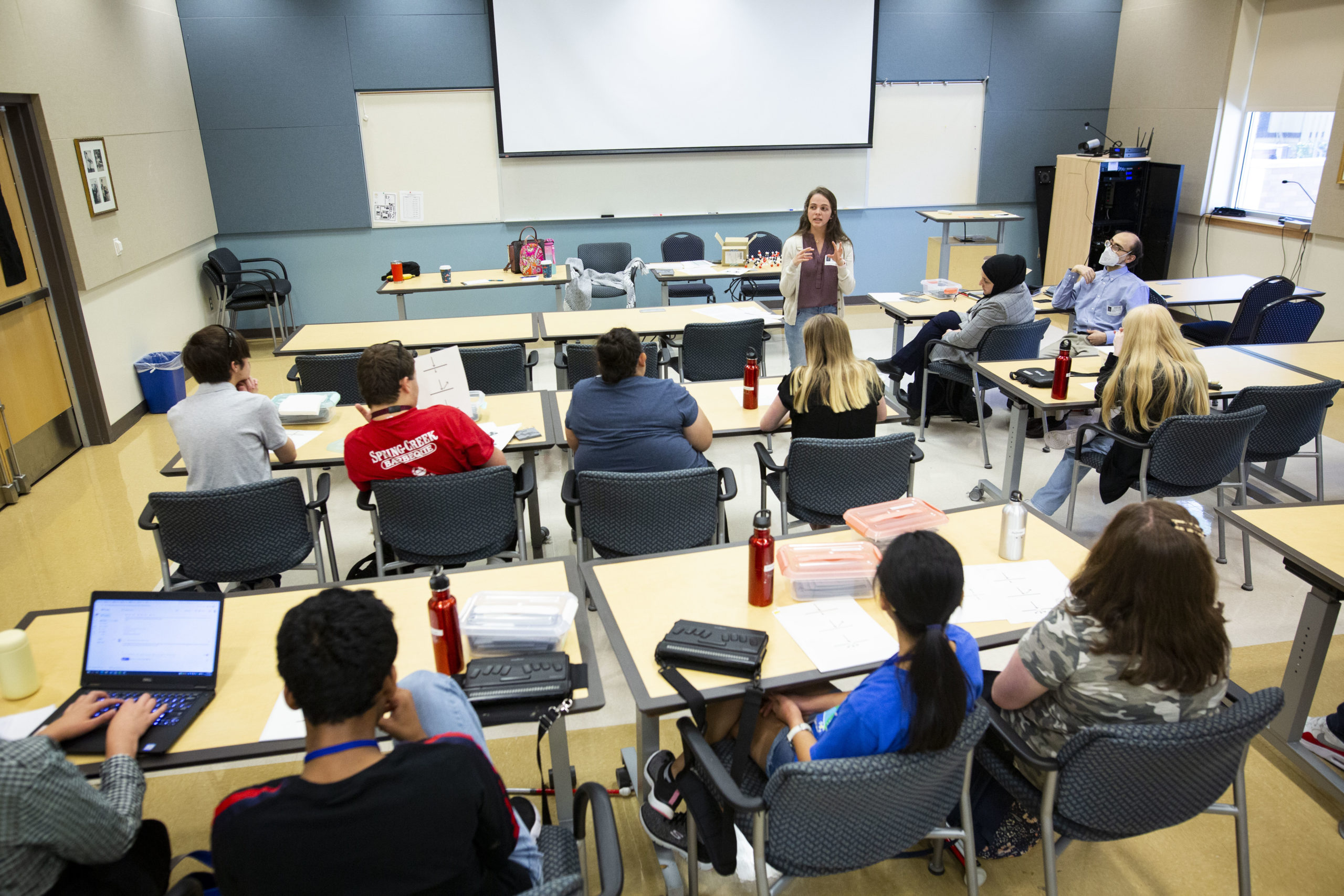 A woman stands in front of a group of students in a classroom. The students are seen in their desks from behind and the woman is facing them, speaking.