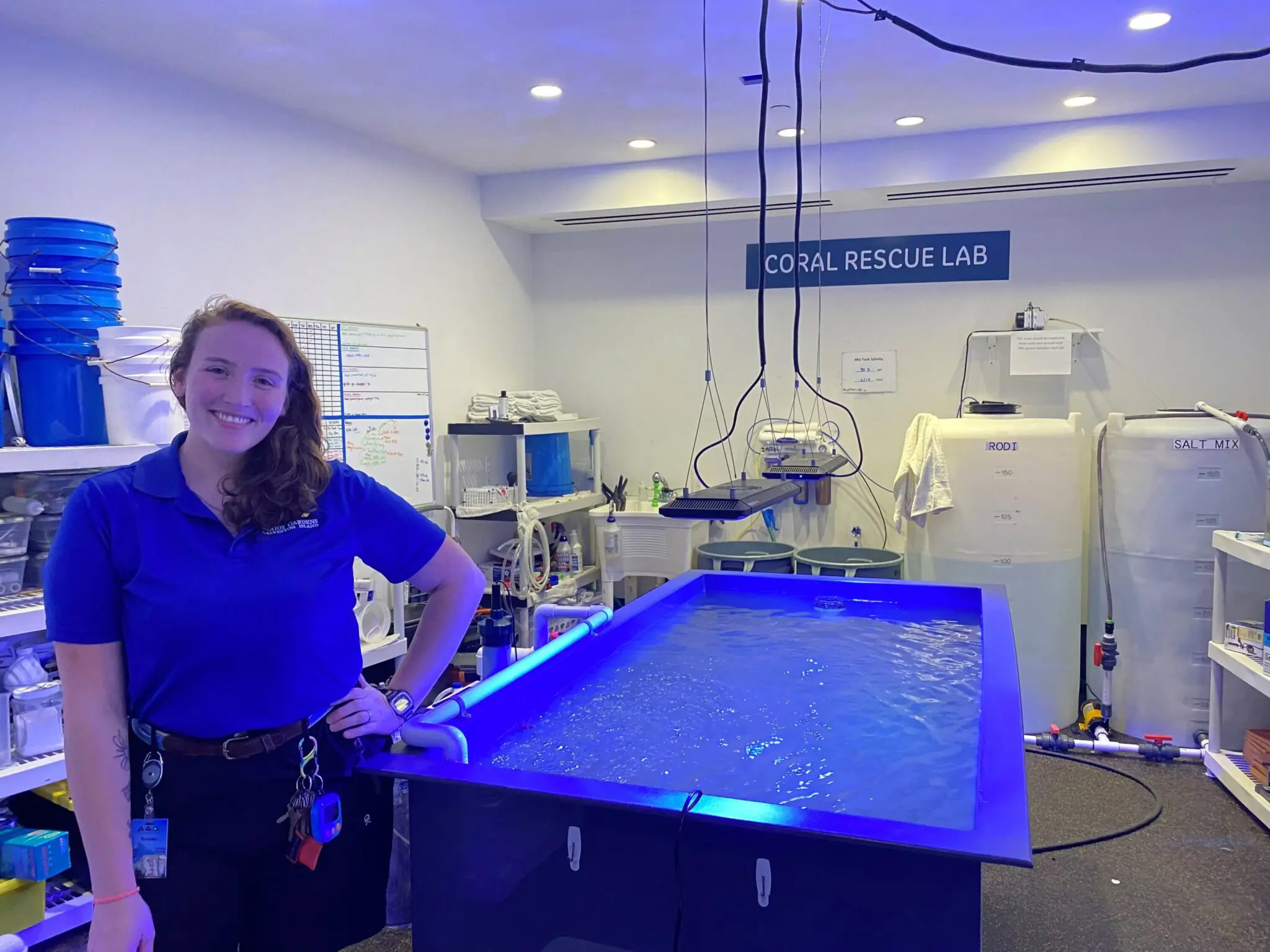 A woman stands smiling next to a large tank filled with water. A sign in the back reads 