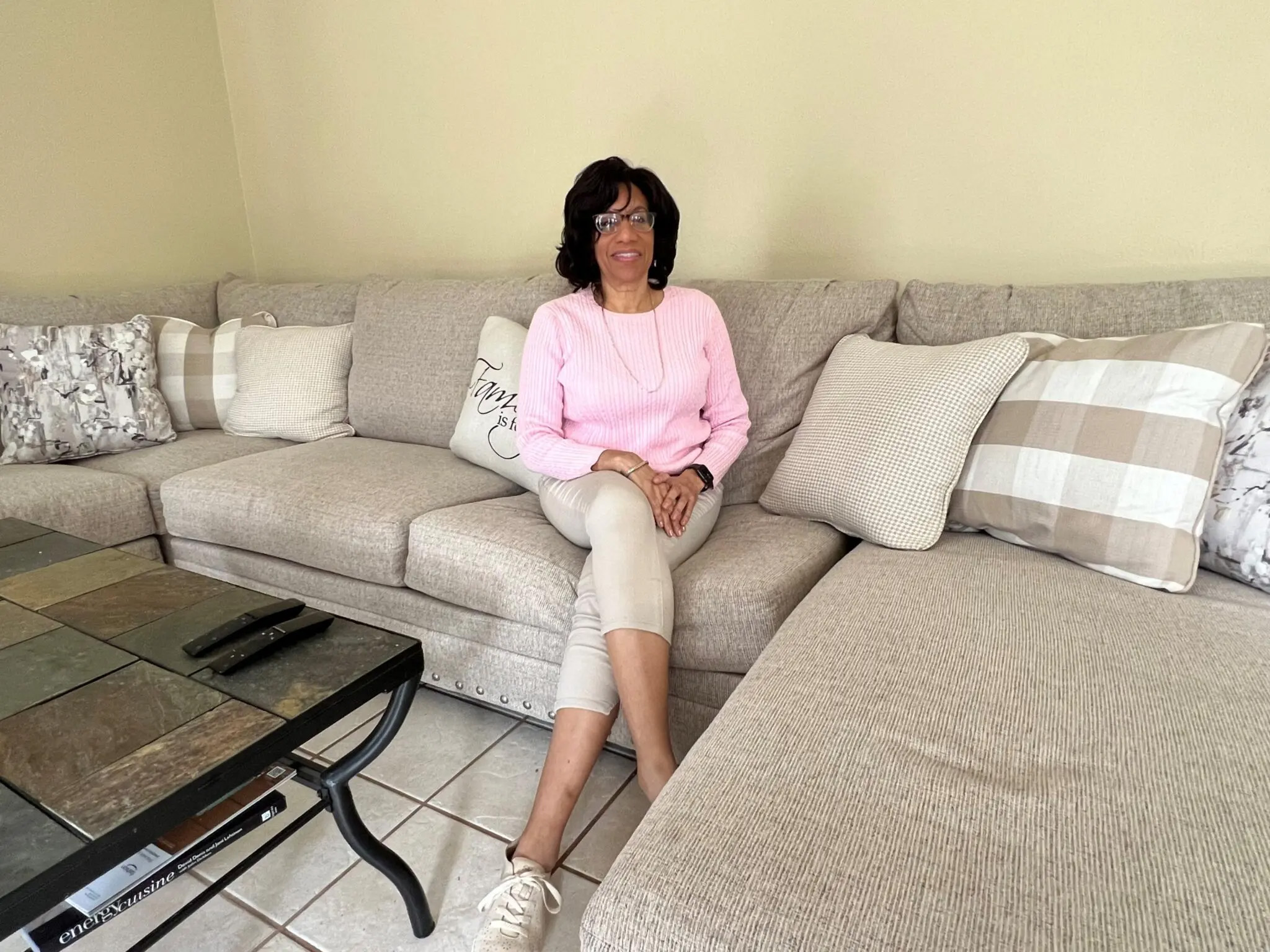 A woman is seated on a sectional sofa facing forward and smiling for the camera. To the side a black, tile-top coffee table is seen. Several pillows of solid or checkered patterns are seen on the sofa. This is Deborah Jackson.