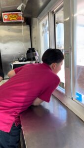 A young, 12-year-old boy is seen from the side looking out the window of his family's food truck.