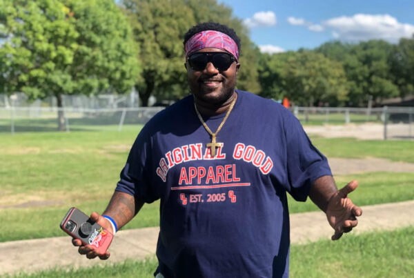 A man is seen from the front, smiling and holding his arms out. In his right hand he holds a phone. He's wearing sunglasses, a bandana around his forehead and a large gold chain with a cross on it. This is Antong Lucky.
