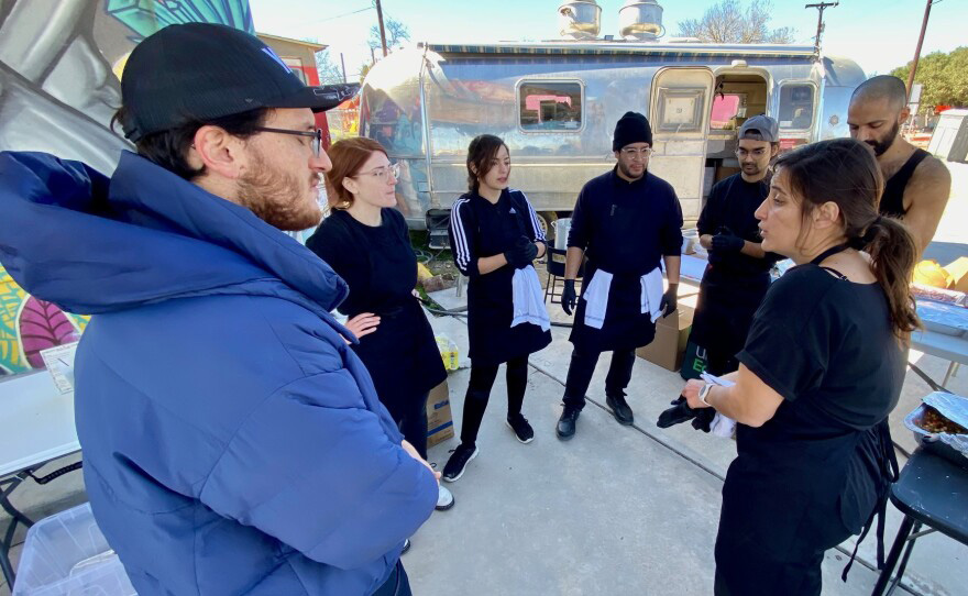 Six people are seen gathered around a woman wearing an apron in the middle of speaking. This is Moureen Kaki, who started the Saha pop-up, and those gathered around are her friends and partners who help run it.