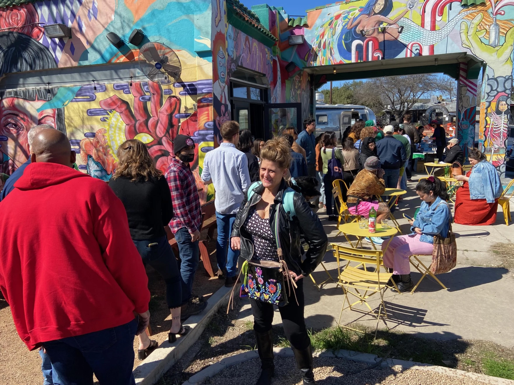 Dozens of people stand in line at the outdoors pop-up for Saha. Next to the line are some tables where customers are already seated. The line winds through a building covered in colorful artworks.