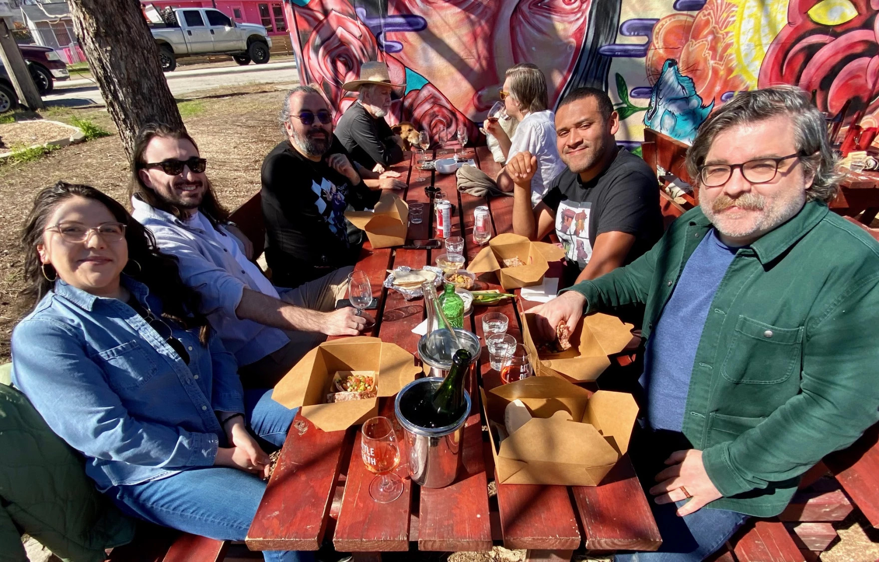 Seven people sit at a table enjoying their meals at the Saha outdoors pop-up. The five in the foreground are looking up from their meals, which sit on the table held in small cardboard containers, and smile at the camera.