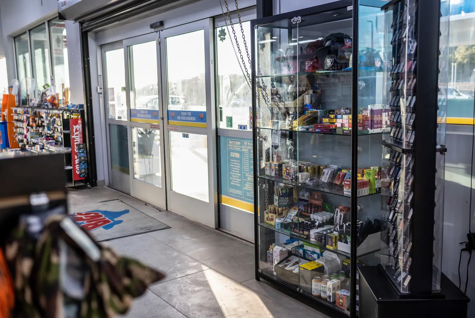 The interior of a convenience store is seen looking towards the sliding doors leading outside. Next to the doors is a tall glass case with dozens of vape products on its shelves.