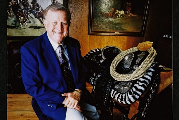 Red McCombs, wearing a blue suit jacket, appears seated next to a saddle