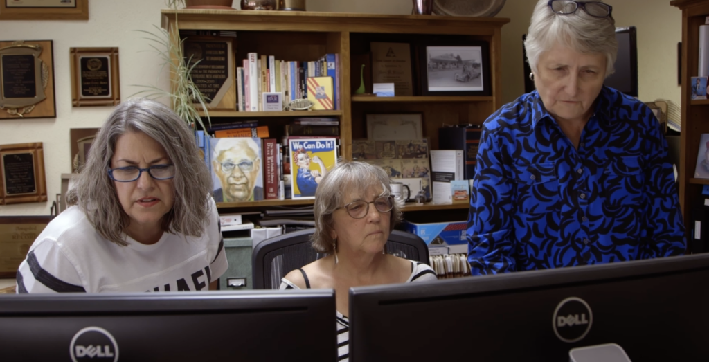 three women sit at two computer screens in an office