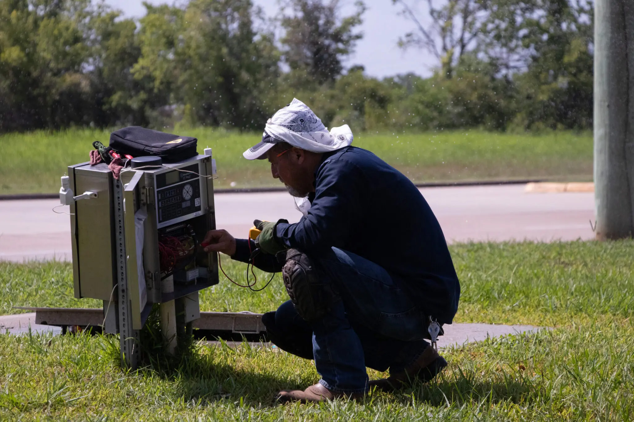 A man crouches while inspecting a sprinkler system box outdoors. He's wearing a hat with a bandana wrapped around for extra sun protection. This is Jose Ruiz.