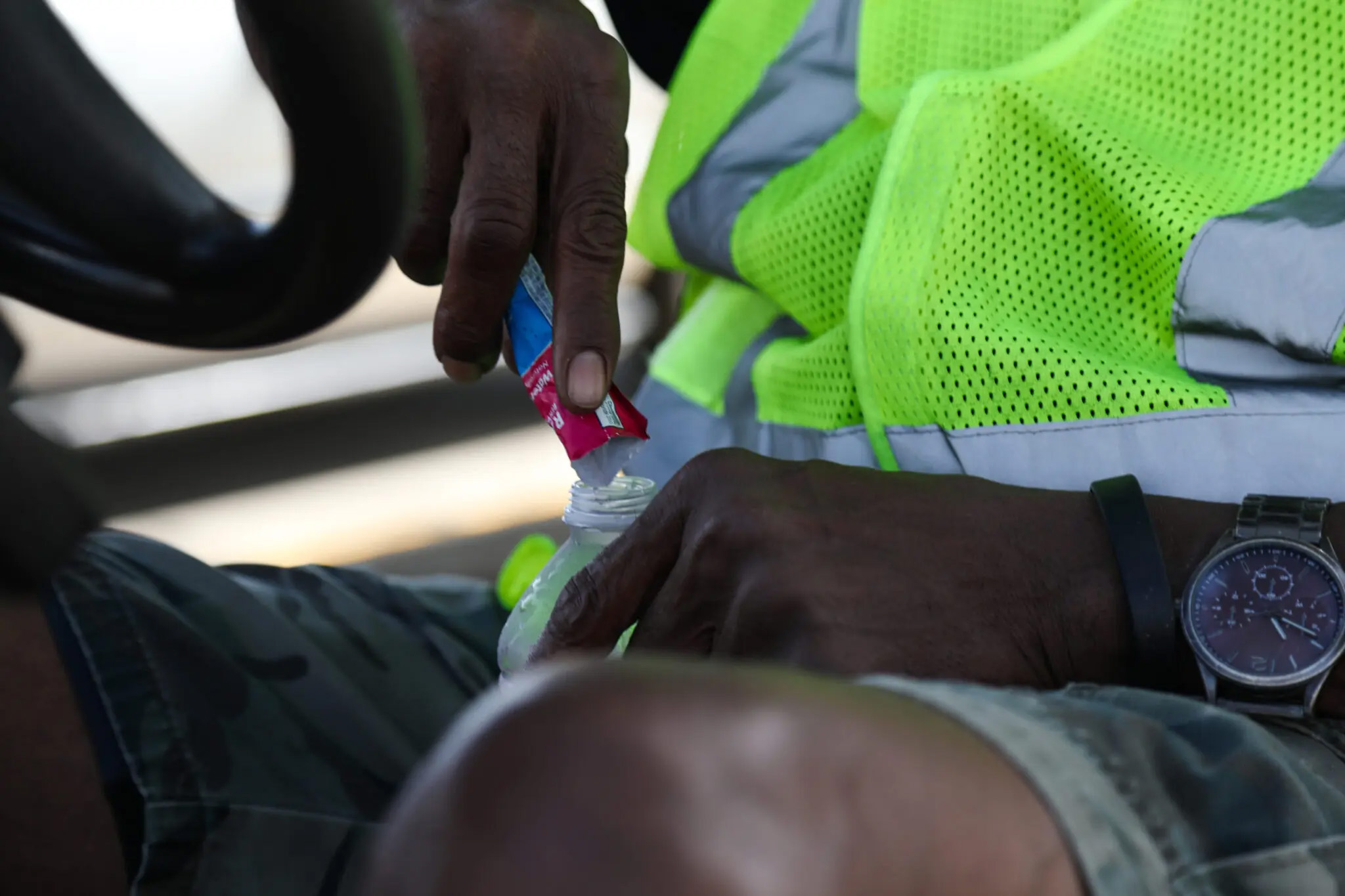 A close-up shot showing the hands of a Walmart associate pouring a small electrolyte packet into his opened plastic water bottle.