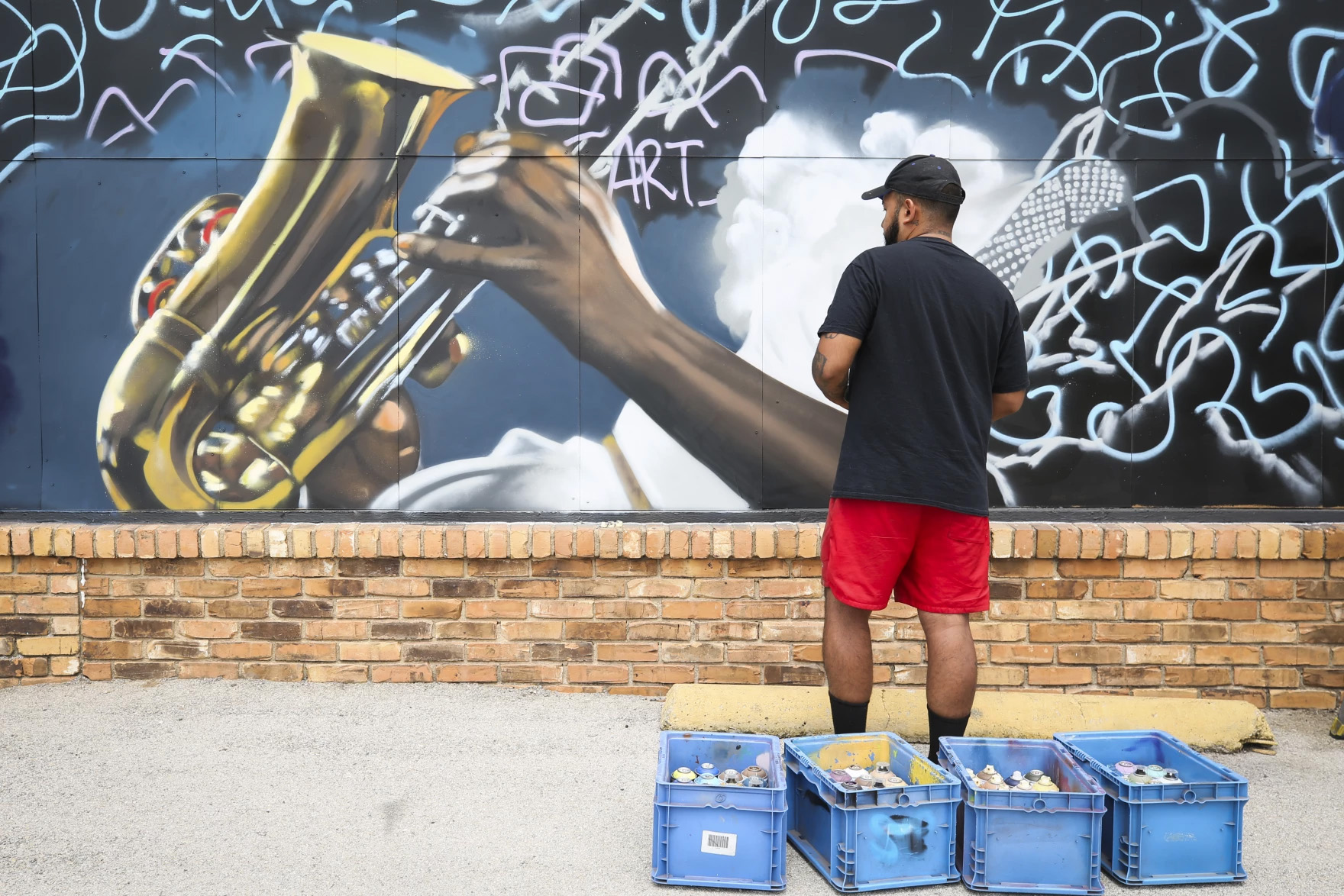 A man stands in front of a mural depicting a saxophone being played surrounded by white squiggle lines on a black backdrop. The man is Juan Velasquez, who is painting the mural.